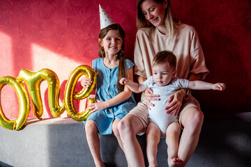 Young mother sits on sofa with small children near red wall on gray sofa. Family with one parent celebrates the first year of baby. Older sister is holding foil balloons ONE. Lifestyle party at home.