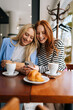© dikushin - Vertical portrait of two cheerful girlfriends women sitting in cafe looking at old photos on smartphone and laughing. Best females friends sitting at coffee shop smiling looking at phone screen.