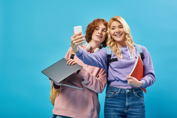 Wall Mural - joyful teenage girl with notebook taking selfie with redhead boyfriend holding laptop on blue