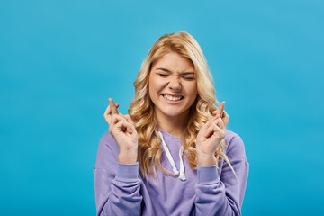 Wall Mural - tense blonde teenage girl with closed eyes holding crossed fingers and wishing luck on blue