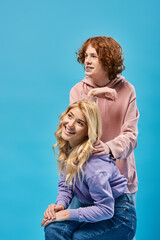 Wall Mural - cheerful redhead guy looking away near blonde girlfriend sitting on blue, happy teenage friends