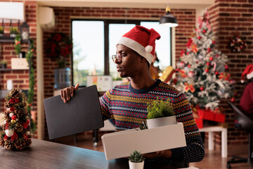 Wall Mural - Upset company worker packing belongings and laptop after being dismissed on christmas eve. Unhappy african american man experiencing unemployment after getting fired during winter holiday