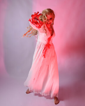 Full length portrait scary vampire zombie bride wearing bloody halloween fantasy costume dress. Dynamic jumping pose gestural arms reaching out. Isolated  studio backdrop with cinematic red lighting.