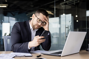 Wall Mural - Upset young man businessman, office worker sitting at desk and holding hand on head, got bad news, reading message