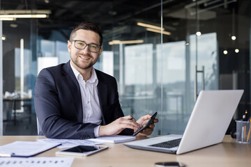 Portrait of a young businessman, accountant, financial analyst working in the office with a calculator and documents, sitting at a desk with a laptop and smiling at the camera