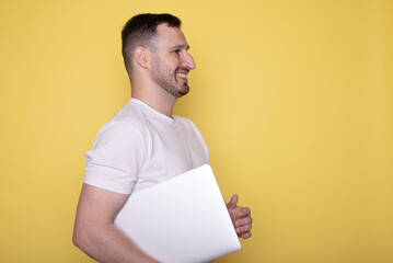 Amazed smiling handsome young bearded man holding underarm laptop pc computer isolated on yellow background.