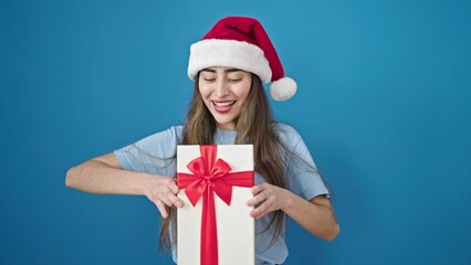 Wall Mural - Young beautiful hispanic woman wearing christmas hat holding gift over isolated blue background