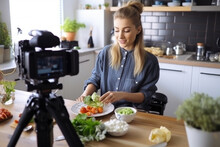 A young woman food blogger cooking salad in front of smartphone camera while recording vlog video and live streaming at home in kitchen.