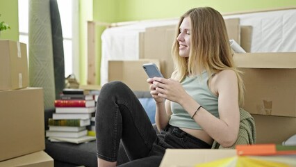 Poster - Young blonde woman using smartphone sitting on sofa at new home