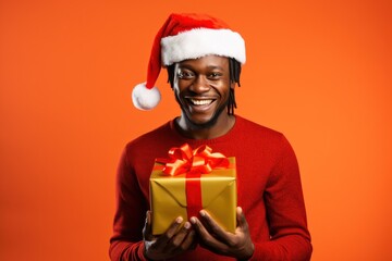 Poster - dark-skinned smiling man with a box with a bow and a Santa Claus hat on an orange background.