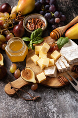 Cheese plate, still life of seasonal wine snacks. Brie cheese, fresh figs, grapes, cheese cubes, pears, pecan nuts and honey on cutting board on dark stone background. Copy space.