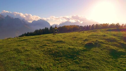 Wall Mural - AERIAL: Stunning alpine views at the hilltop enjoyed by a woman with her dog. Relaxed sporty lady sits on electric bike and admires last rays of autumn sun illuminating beautiful mountain landscape.