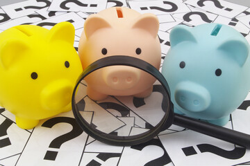 Piggy banks under magnifying glass on question marks background.	