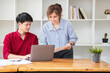 © PHAISITSAWAN - Cheerful business people using a laptop in an office. Happy young entrepreneurs smiling while working together in a modern workspace. Two young business people are sitting together at a table.