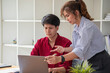© PHAISITSAWAN - Cheerful business people using a laptop in an office. Happy young entrepreneurs smiling while working together in a modern workspace. Two young business people are sitting together at a table.