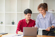 © PHAISITSAWAN - Cheerful business people using a laptop in an office. Happy young entrepreneurs smiling while working together in a modern workspace. Two young business people are sitting together at a table.