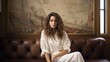 © Romana - a photo of gorgeous young caucasian woman sitting on a couch in a luxurious posh living room, parisian style interior with tall windows, white paneled walls, fireplace, golden sophisticated decoration
