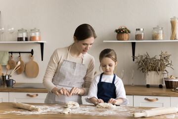 Caring mom teaching cute toddler girl in apron to cook traditional homemade pastry, baking homemade dessert, beating raw sticky dough on floury table, enjoying motherhood, culinary hobby