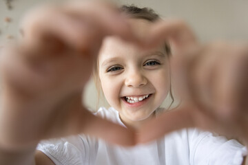 Positive sweet little preschool girl looking at camera through hand heart frame with toothy smile. Cheerful pretty little kid joining fingers, showing gesture sign of happiness, love, happy childhood