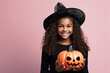 niña vestida de bruja con sobrero en negro sosteniendo entre las manos una calabaza naranja con cara de terror sobre fondo rosa, concepto halloween