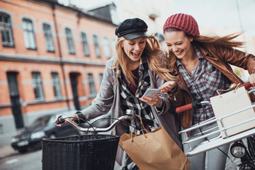 Wall Mural - Young lesbian couple using a smartphone and riding bikes together while shopping in the city