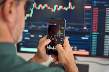 Investor trader broker analyzing financial crypto stock trade market on smartphone risk digital price data in mobile app buying bank shares, doing investment strategy analysis on phone. Over shoulder.