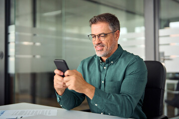 Busy smiling business man executive of mid age looking at smartphone digital technology, older mature businessman entrepreneur investor using cell phone working on mobile cellphone in office.