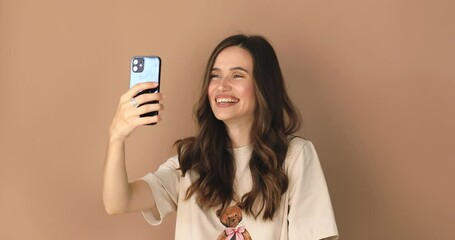 Wall Mural -  Happy young woman blogger influencer holding modern smart phone wave hand hello. Smiling vlogger girl looking at mobile make video call, shooting vlog taking selfie on beige background, hi gesture.