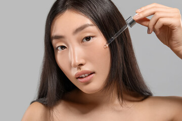 Wall Mural - Beautiful young woman applying cosmetic serum onto her face on grey background, closeup