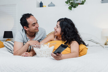 Wall Mural - Latin couple watching a movie lying on bed at home in Mexico Latin America, hispanic people eating popcorn on bedroom