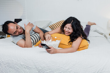 Wall Mural - Latin couple watching a movie lying on bed at home in Mexico Latin America, hispanic people eating popcorn on bedroom