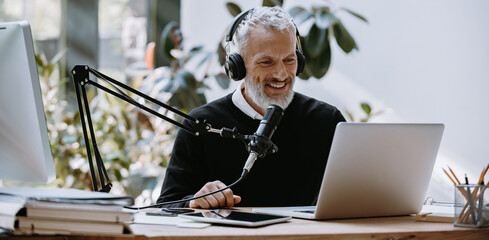 Cheerful mature podcaster in headphones using microphone while broadcasting from studio