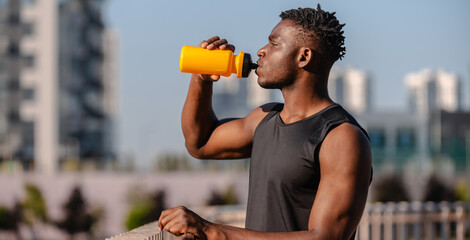 Canvas Print - Handsome African man in sportswear drinking water while resting after training outdoors