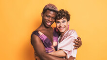 two drag men couple in make up embracing together looking to camera on yellow