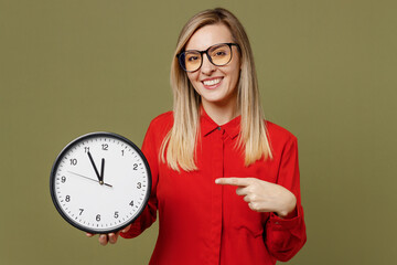 Wall Mural - Young smiling happy fun cheerful woman she wearing red shirt casual clothes glasses holding in hands point finger on clock looking camera isolated on plain pastel green background. Lifestyle concept.