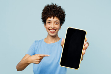 Wall Mural - Young woman of African American ethnicity in t-shirt casual clothes hold in hand use point finger on mobile cell phone with blank screen workspace area isolated on plain pastel light blue background