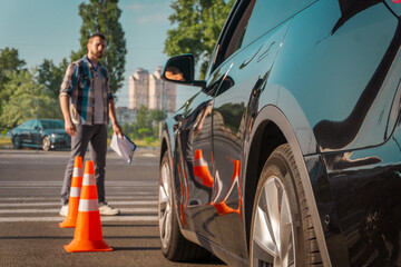 Male instructor clipboard in his hand while looking at how lady parking in the driving school outdoors. Traffic cones near car. Driving test, driver courses, exam concept