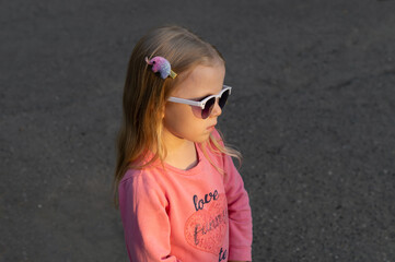 Portrait beautiful little girl with blond hair in sunglasses standing on the street on a background of gray asphalt in the sunlight