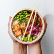 Hands holding a poke bowl with salmon raw fish, rice, edamame beans with chopsticks, close up of poke on white background, top table view photograph
