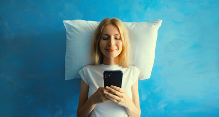 Wall Mural - Healthy sleep, happy relaxed sleepy young woman with mobile phone on white soft comfortable pillow imagining she lying on the bed on blue sky with clouds background