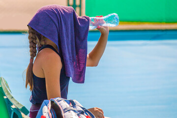 Wall Mural - A young girl tennis player is resting while sitting on a bench in between games and drinks water