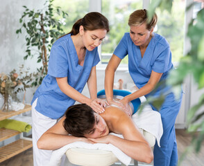 Massage in four hands. Two female masseuses perform stroking and rubbing of muscles of back and shoulders to young guy client of spa salon. Restorative relaxing therapeutic procedures