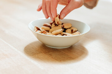 Wall Mural - Brazil nuts in the diet. Healthy fats in the diet. Brazil nuts in a white ceramic cup in hand on a wooden table.hand takes brazil nuts from a plate close-up.