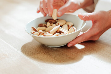 Wall Mural - Brazil nuts in the diet. Healthy fats in the diet. Brazil nuts in a white ceramic cup in hand on a wooden table.hand takes brazil nuts from a plate close-up.Nuts and seeds.Useful healthy snack. 