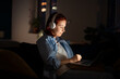 Focused freelancer woman in headphones sitting on sofa, working on laptop at home late night. Side view girl with red hair watching media on computer, wear headset listen music in dark room. 