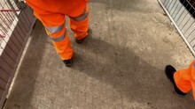 POV Of Group Of Feet Walking Wearing Bright Orange High Visibility Trousers At Construction Site