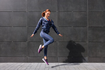 Poster - Beautiful woman in gym clothes jumping near dark grey wall on street, space for text