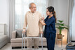 Front view of a happy senior Asian man was standing using a walker with the assistance of a young Asian female nurse in a blue uniform was supporting his arm and giving him a thumbs up in living room.