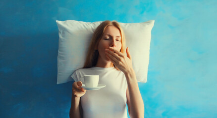 Wall Mural - Lazy woman waking up after sleeping and yawning on white soft comfortable pillow imagining she lying on the bed holds cup of coffee on blue background at home