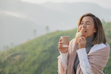 Wall Mural - Relaxed woman breathing fresh air holding coffee cup and croissant for breakfast in autumn in a forest sitting on balcony and looking at mountains and green nature.Trekking concept..